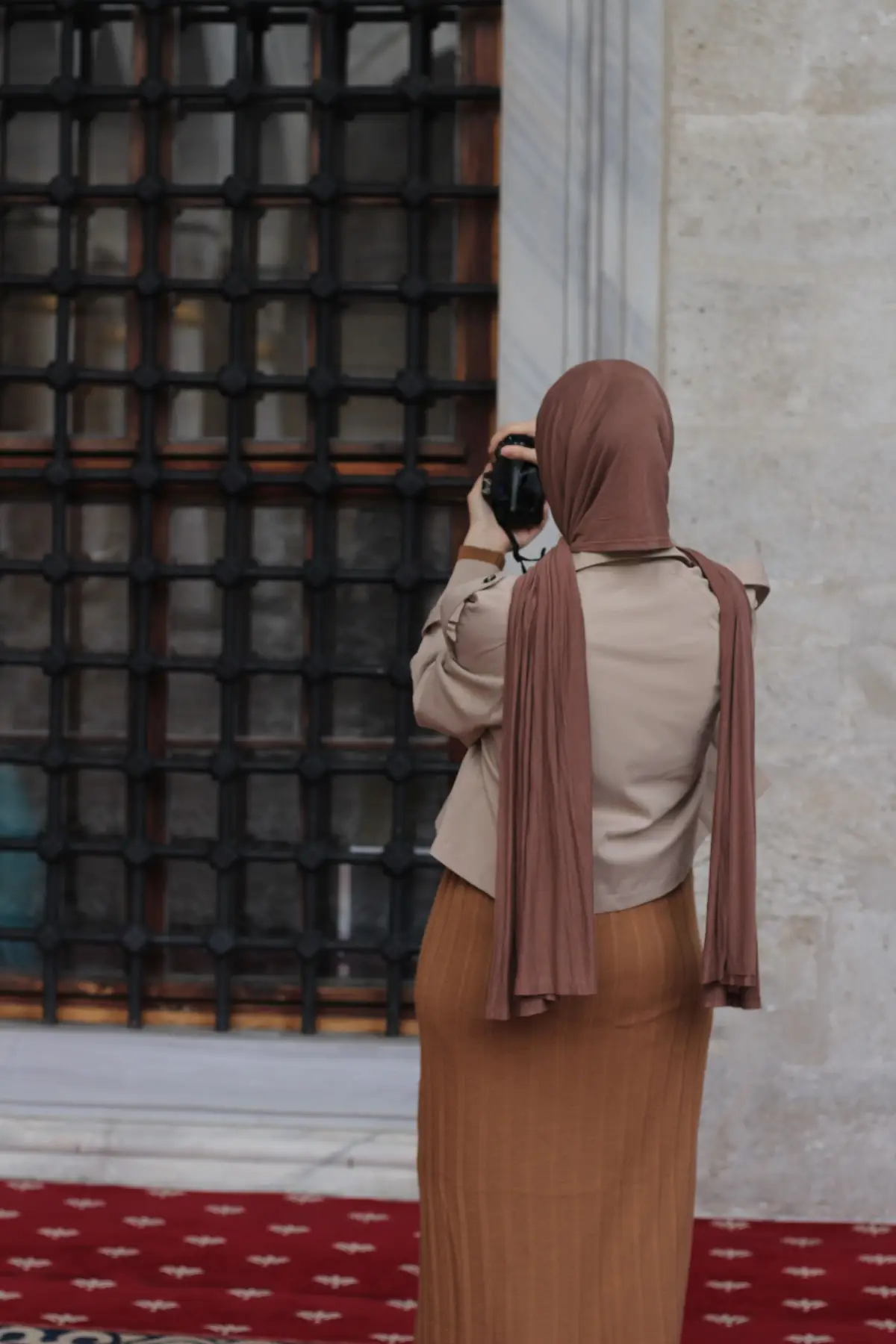 A woman in a beige hijab and camel skirt photographing an old building